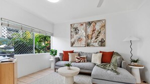 Una sala de estar moderna con un sofá gris, una mesa de centro y un televisor sobre un mueble de madera. Hay dos cuadros colgados en la pared y una planta en maceta sobre una mesita auxiliar.