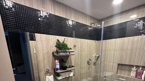 Un baño moderno con cabina de ducha de vidrio, inodoro y lavamanos. Hay estantes con artículos de aseo y una planta en maceta.