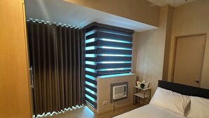A hotel room with a bed, a desk, a chair, a window with blinds, a radiator, and a door.