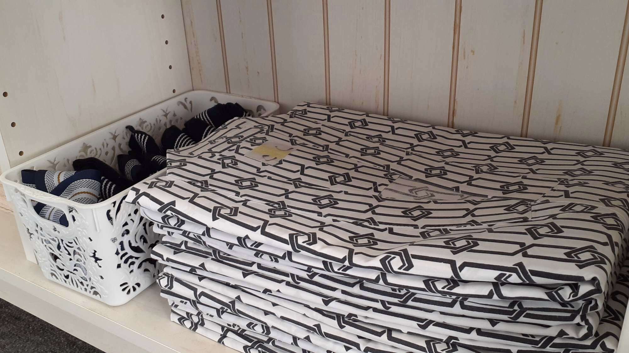 Un lit dont la literie est soigneusement pliée, recouvert d’une couvre-lit à motifs géométriques, un panier décoratif blanc contenant des serviettes pliées, et un mur blanc orné de lignes verticales.