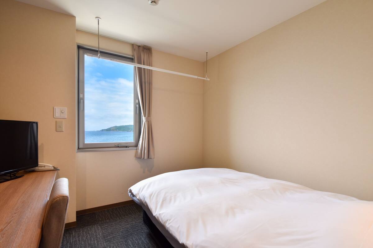 A hotel room with a bed, a desk with a television, a window with curtains, and a view of the sea.