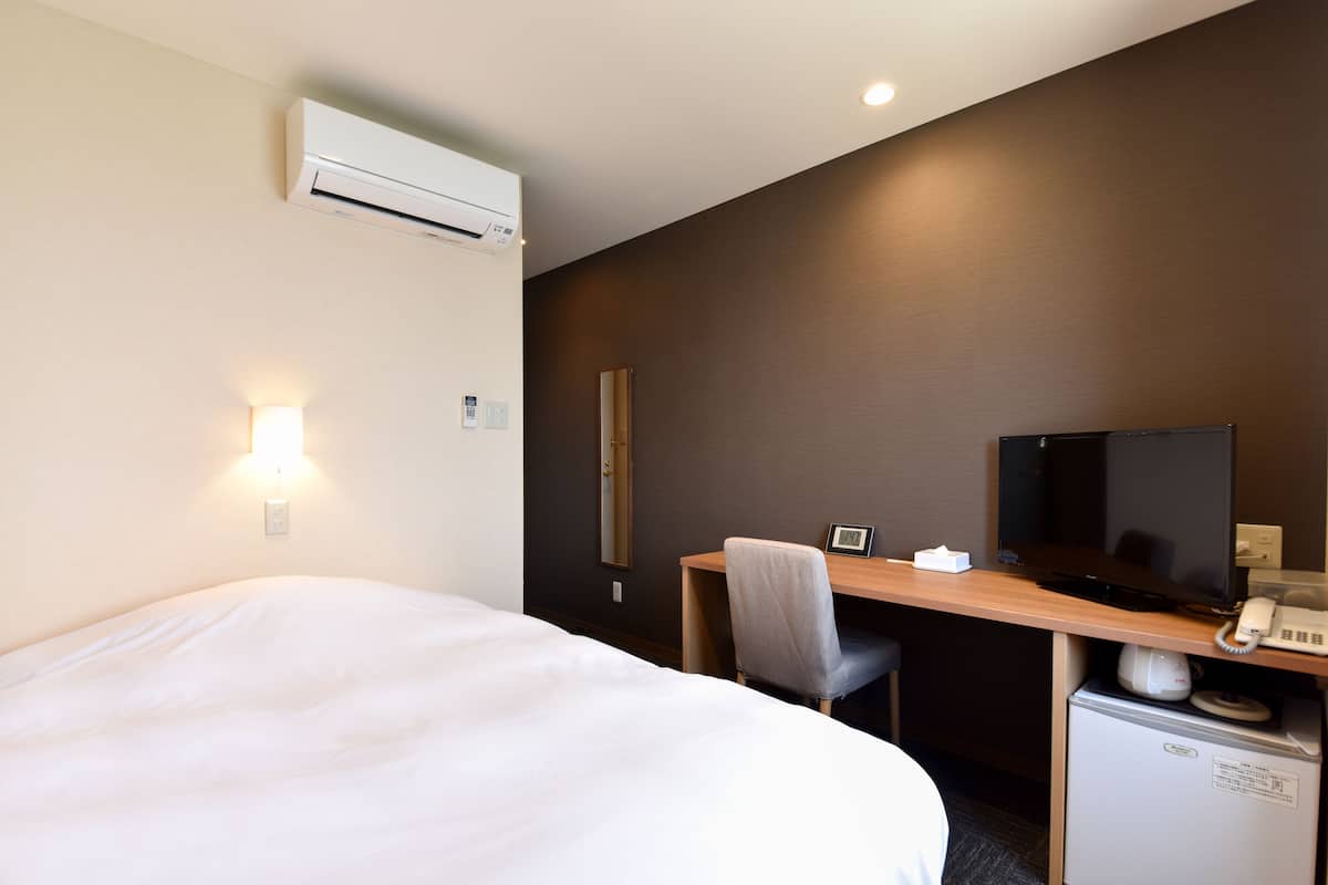 A hotel room with a bed, a desk with a television, a small refrigerator, and an air conditioning unit.