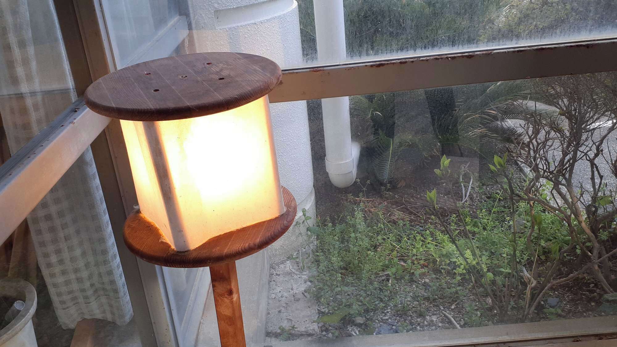 A window with a wooden lantern hanging on the sill, a white window frame, and a view of a garden with various plants and a white pipe.