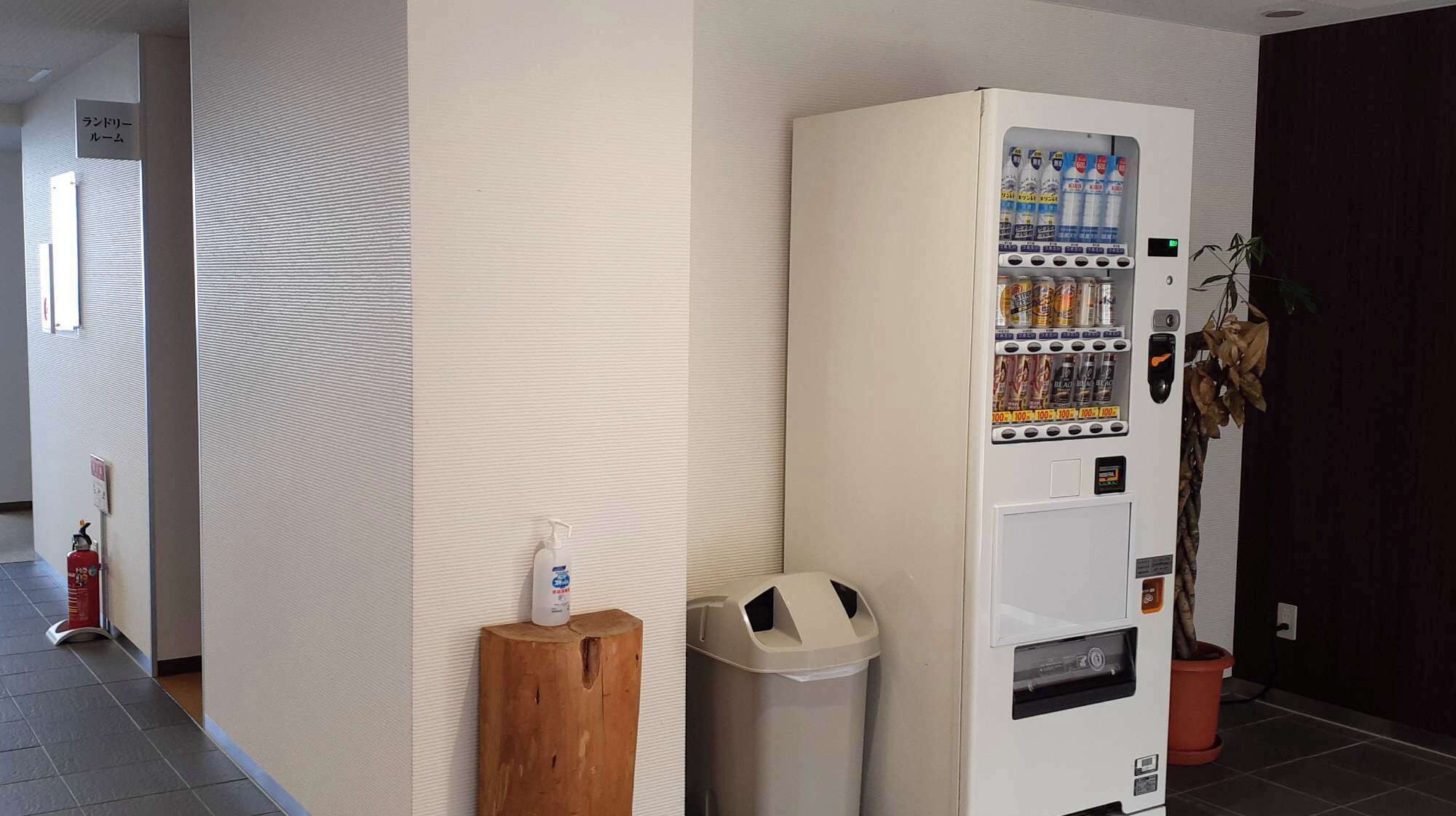 Una máquina expendedora con bebidas y aperitivos, un cubo de basura, un taburete pequeño de madera y una planta en maceta.
