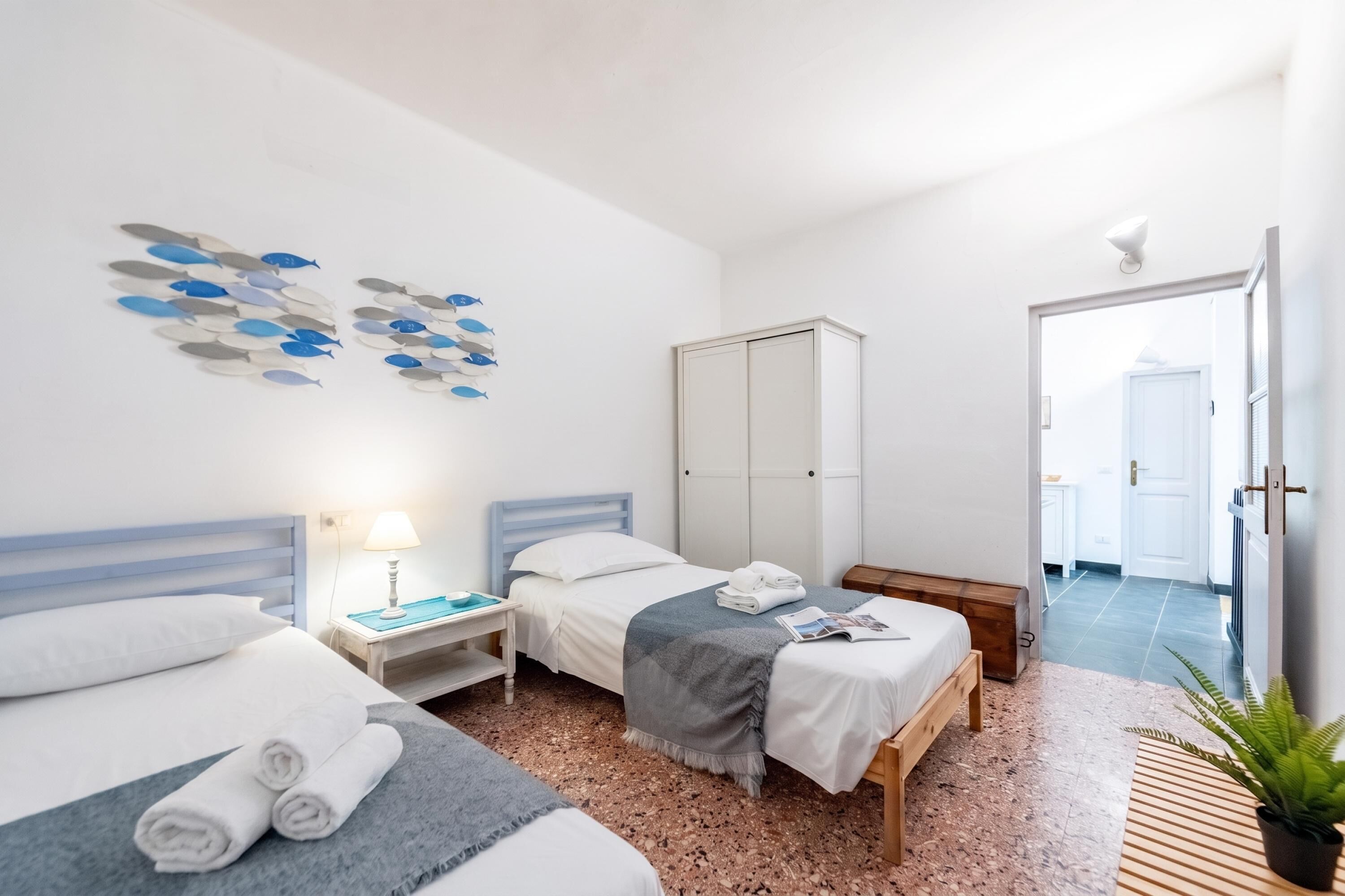 A hotel room with two beds, a nightstand with a lamp, a wardrobe, and a view of the hallway.