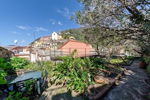 A courtyard with a table and chairs, surrounded by greenery and a view of a hillside with buildings.