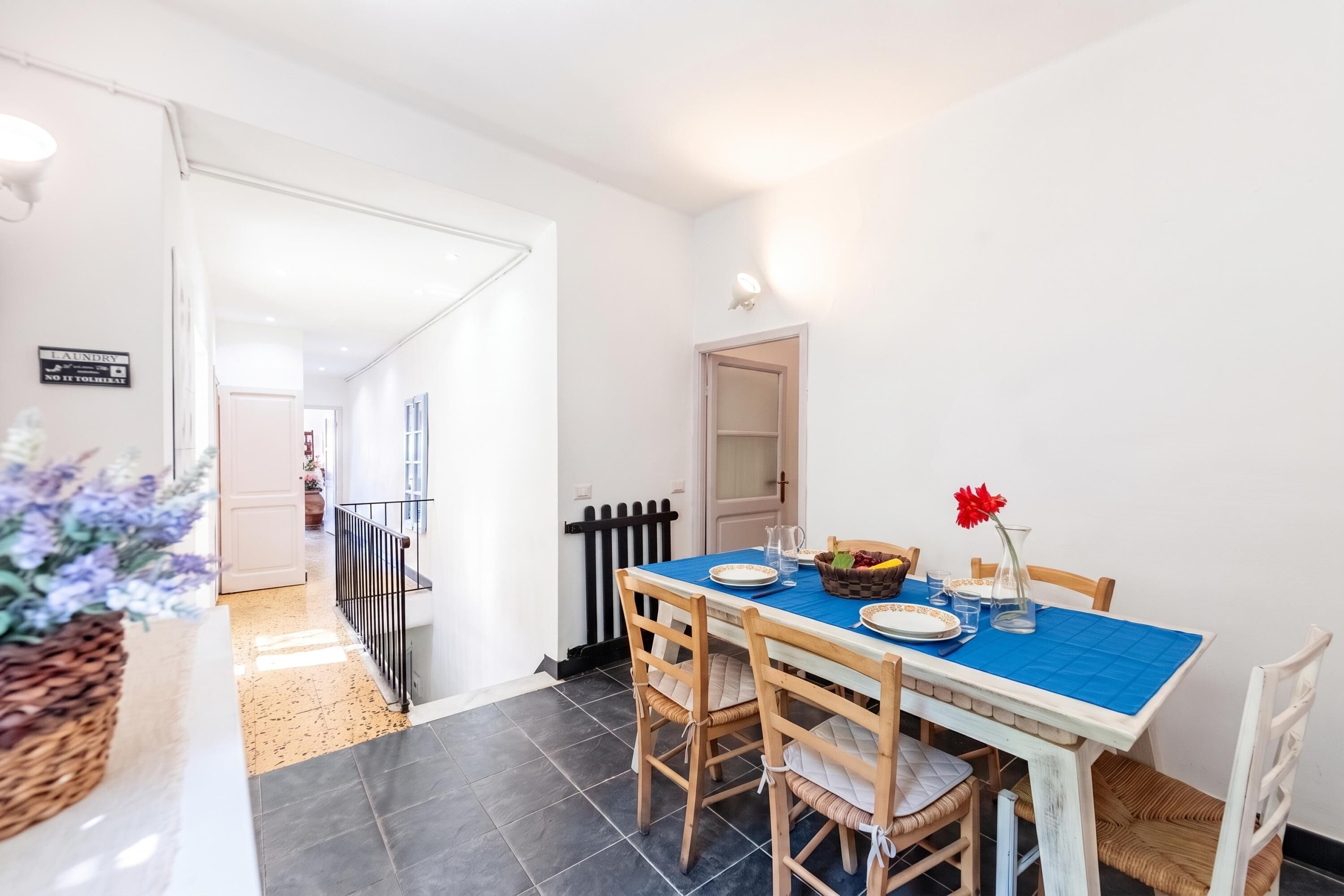 A dining area with a table set for two, a basket of fruit, and a vase with a red flower.
