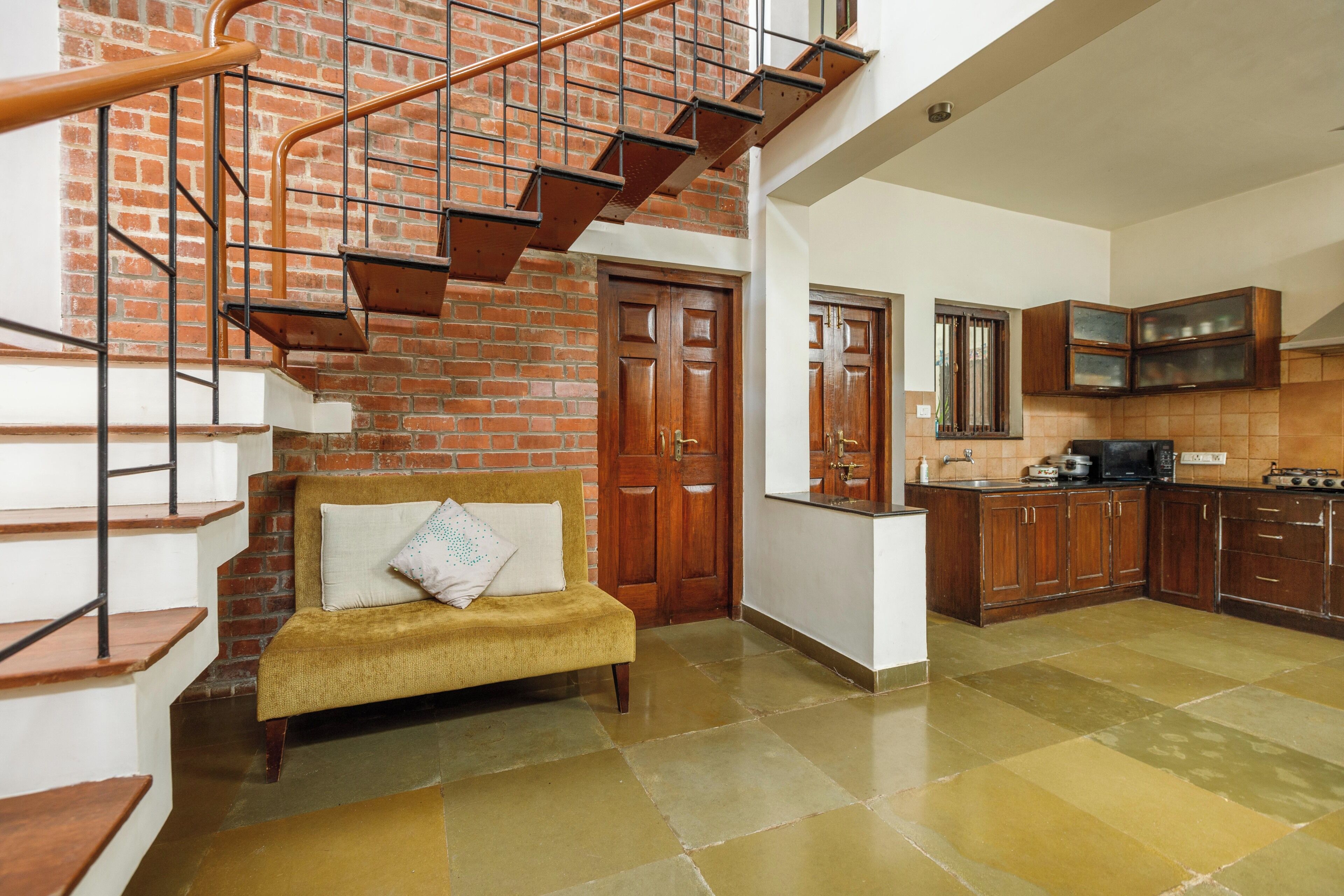 A staircase with a metal railing leading to a wooden door, a sofa with cushions, and a kitchen area with wooden cabinets and a countertop.