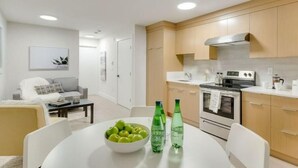 Een moderne keuken met houten kasten, een roestvrijstalen afzuigkap en een wit aanrecht. Er staat een eettafel met een schaal groene appels en twee flessen bruisend water.