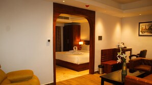 Habitación de hotel con piso de madera, una cama con edredón blanco, una mesita de madera, un sofá de cuero marrón y una pequeña mesa con un florero.
