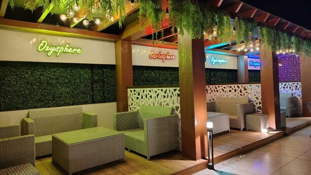 Un área de estar al aire libre con mobiliario de mimbre y cojines verdes, bajo una pérgola de madera con plantas colgantes.