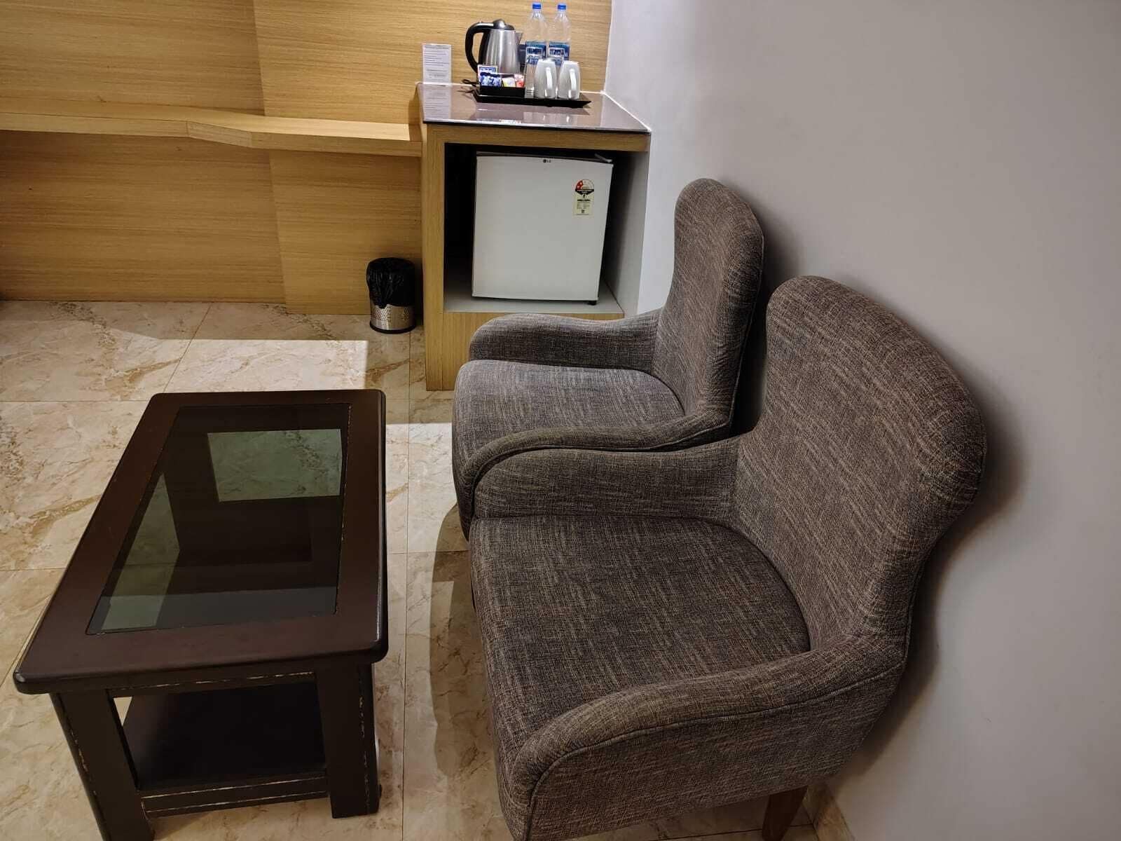 A small room with a wooden wall, a glass-top table, two grey upholstered chairs, a small side table with a kettle and bottles, and a white refrigerator.