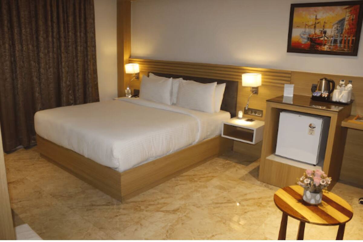 A hotel room with a large bed, a nightstand, a small refrigerator, and a wooden side table with a vase of flowers.
