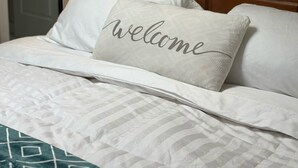 A neatly made bed with a "welcome" pillow, a wooden headboard, and a wall-mounted light fixture.