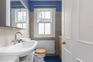 Une salle de bain avec un lavabo blanc, des toilettes en bois, une fenêtre donnant sur un chantier de construction et un radiateur blanc.