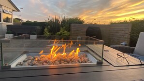 Un foyer extérieur doté d’un plateau en verre, avec des bûches qui brûlent, entouré d’une terrasse en bois et d’un mur en briques.