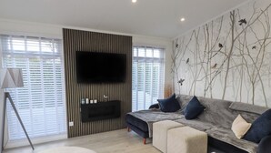 Un salon moderne avec un téléviseur à écran plat fixé sur un mur recouvert de panneaux en bois, un canapé gris agrémenté de coussins bleus, un fauteuil beige et une table basse blanche.
