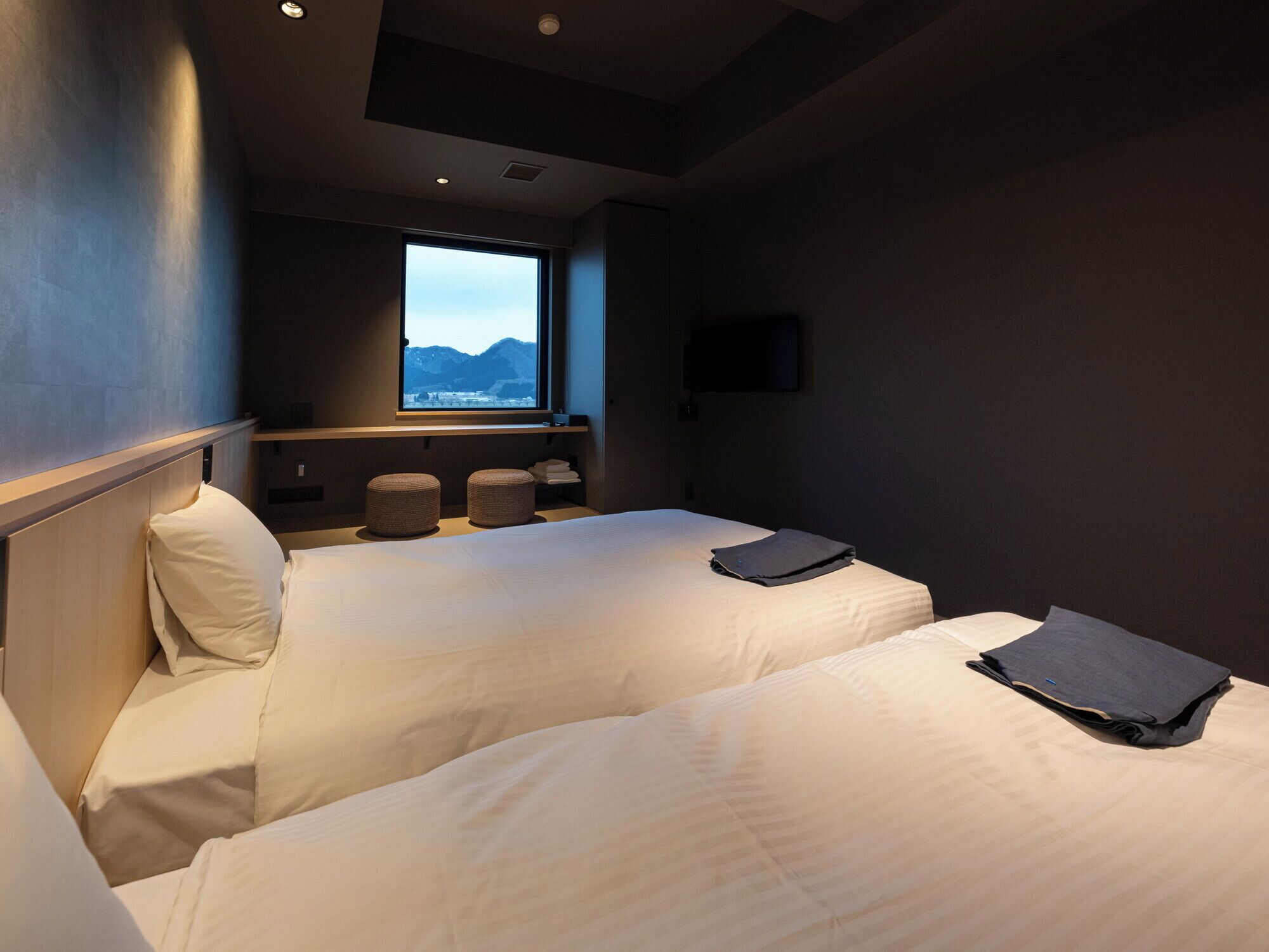 Ein modernes Hotelzimmer mit zwei Betten, einem großen Fenster mit Bergblick, einem Flachbildfernseher und einem kleinen Regal mit Handtüchern.