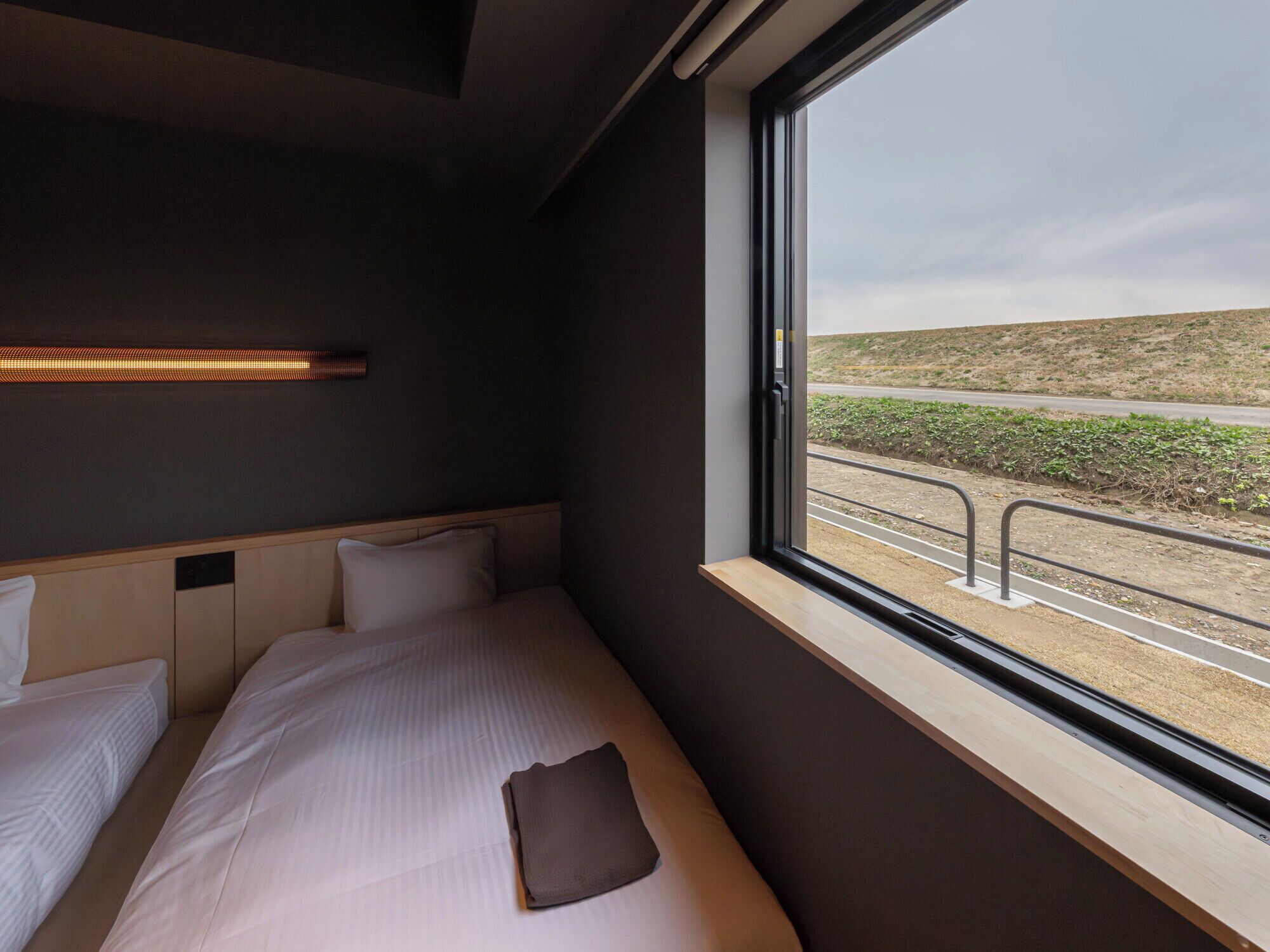 Una cama individual con cabecera, una mesita de noche y una ventana con vista a una carretera y paisaje.
