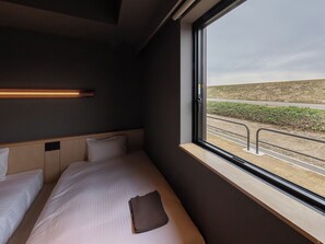Un lit simple avec une tête de lit, une table de chevet et une fenêtre donnant sur une route et un paysage.