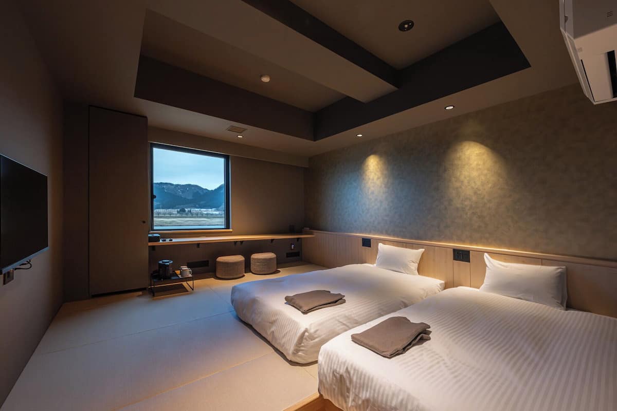 A modern hotel room with two beds, a large window with a mountain view, and a flat-screen TV mounted on the wall.