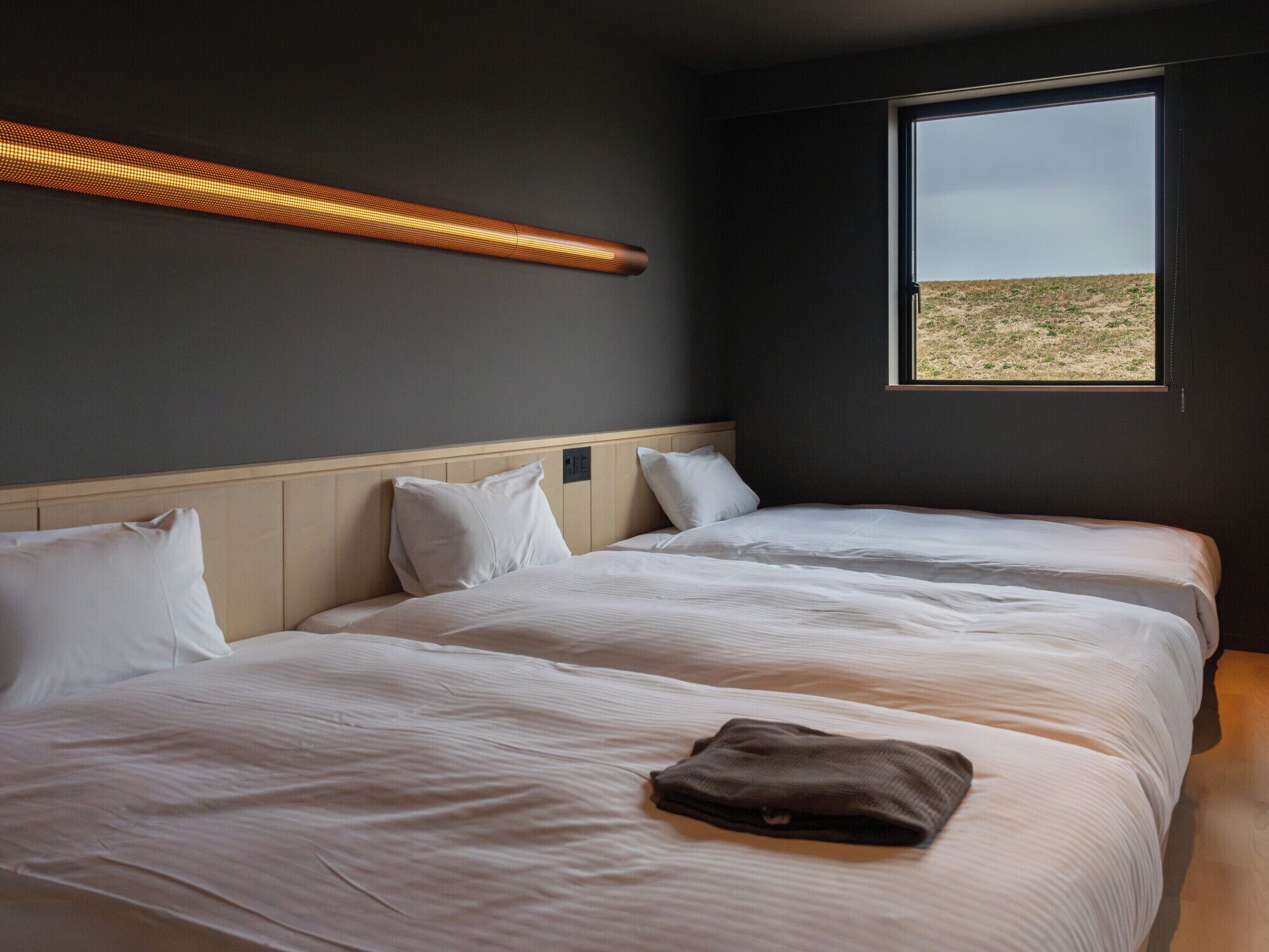 A room with two beds, a window with a view of a field, and a wall-mounted light fixture.