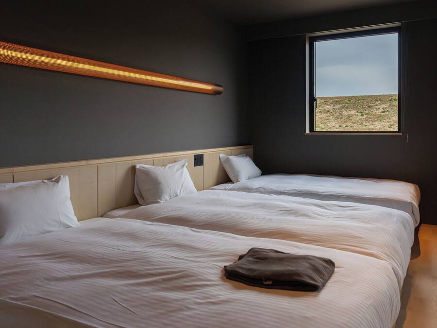 A room with two beds, a window with a view of a field, and a wall-mounted light fixture.