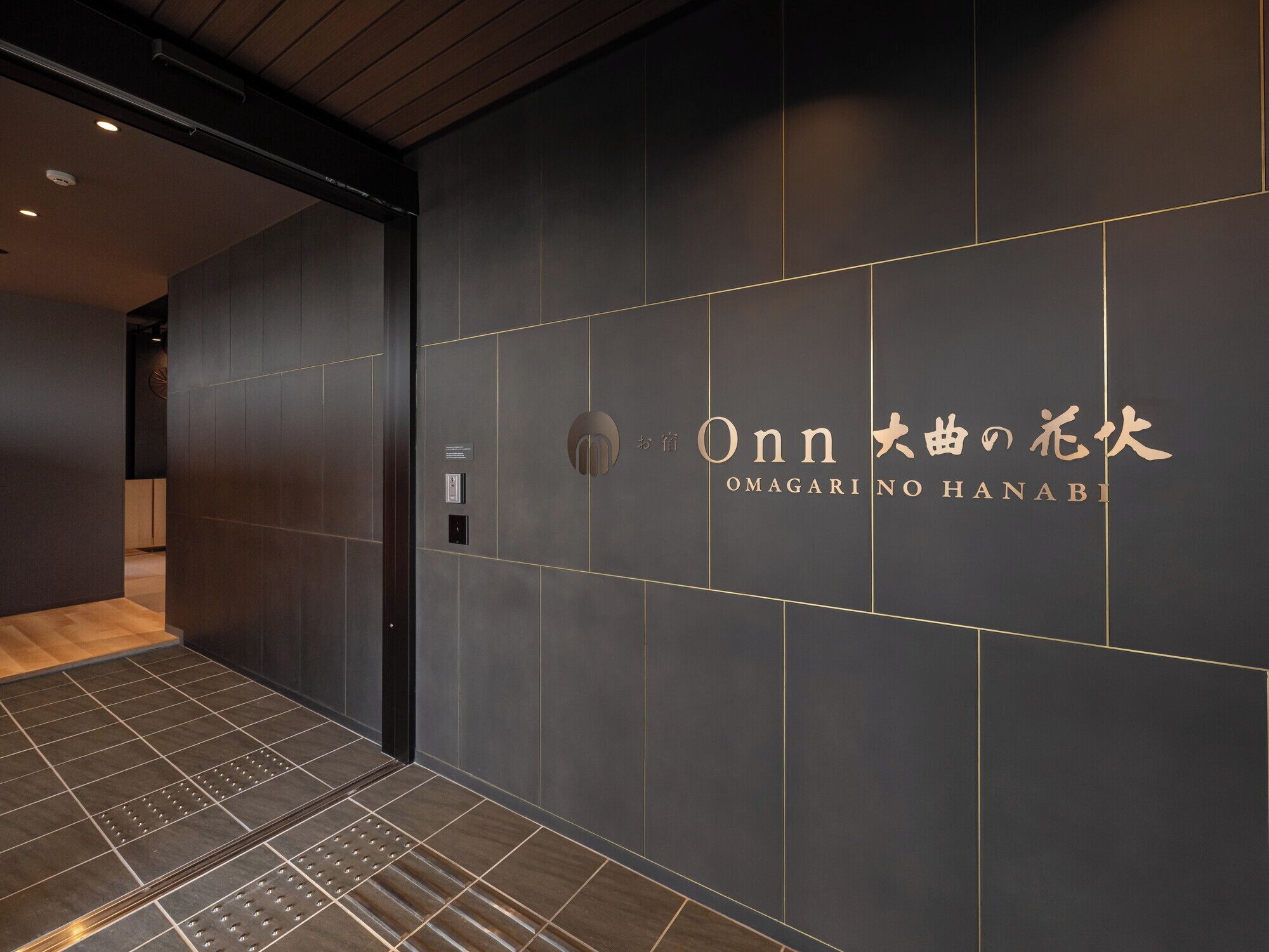 A modern interior with a dark wall and gold accents, featuring a sign that reads "Onn Omagari No Hanabi".