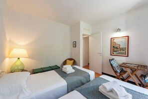 A hotel room with two beds, a green lamp, a painting, and a wooden table.