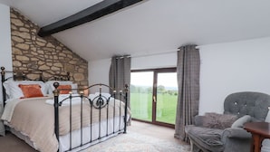A bedroom with a stone wall, a bed with a metal frame, a grey armchair, and a view of a green field through a glass door. - Stoneycross Cottage (Preston)