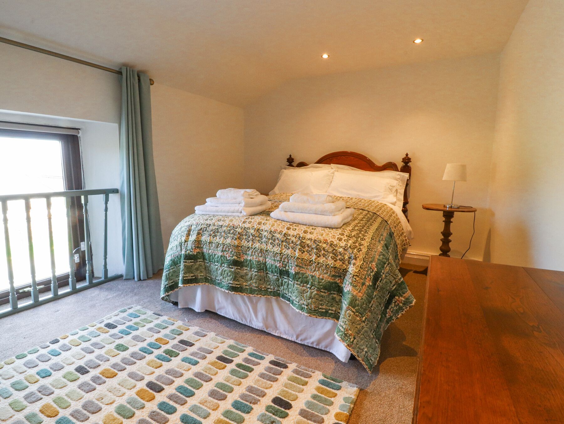 A bedroom with a bed, a bedside table with a lamp, a patterned rug, and a view of the outdoors through a window.