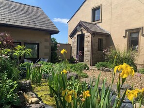 Un jardin agrémenté de fleurs jaunes et un sentier de pierres menant à une maison à la façade en pierre, avec une petite dépendance attenante.