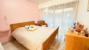 A bedroom with a bed, a wicker chair, a wooden dresser with toiletries, and a window with sheer curtains.