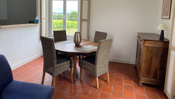 Un coin repas comprenant une table, des chaises en osier, un vase, une lampe et une vue sur la verdure par une fenêtre.