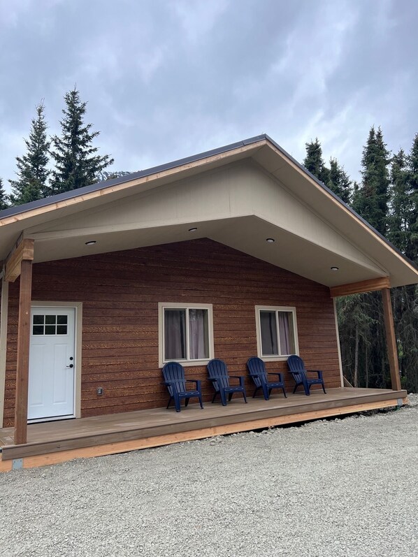 A wooden cabin with a porch, three Adirondack chairs, and a gravel driveway.