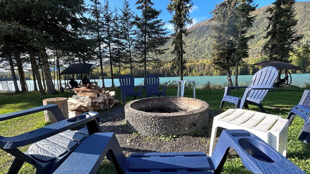An outdoor seating area with Adirondack chairs, a fire pit, and a view of a lake and forest.