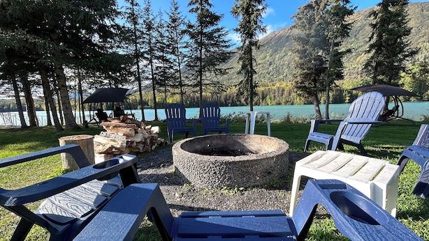 An outdoor seating area with Adirondack chairs, a fire pit, and a view of a lake and forest.