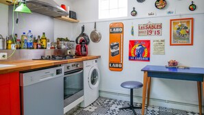 A compact kitchen with a washing machine, a stove, and a sink. There are various kitchen items on the counter, and a table with a chair. The walls are decorated with plates and a poster. - Large apartment tastefully decorated for 5 people (Les Sables-d´Olonne)
