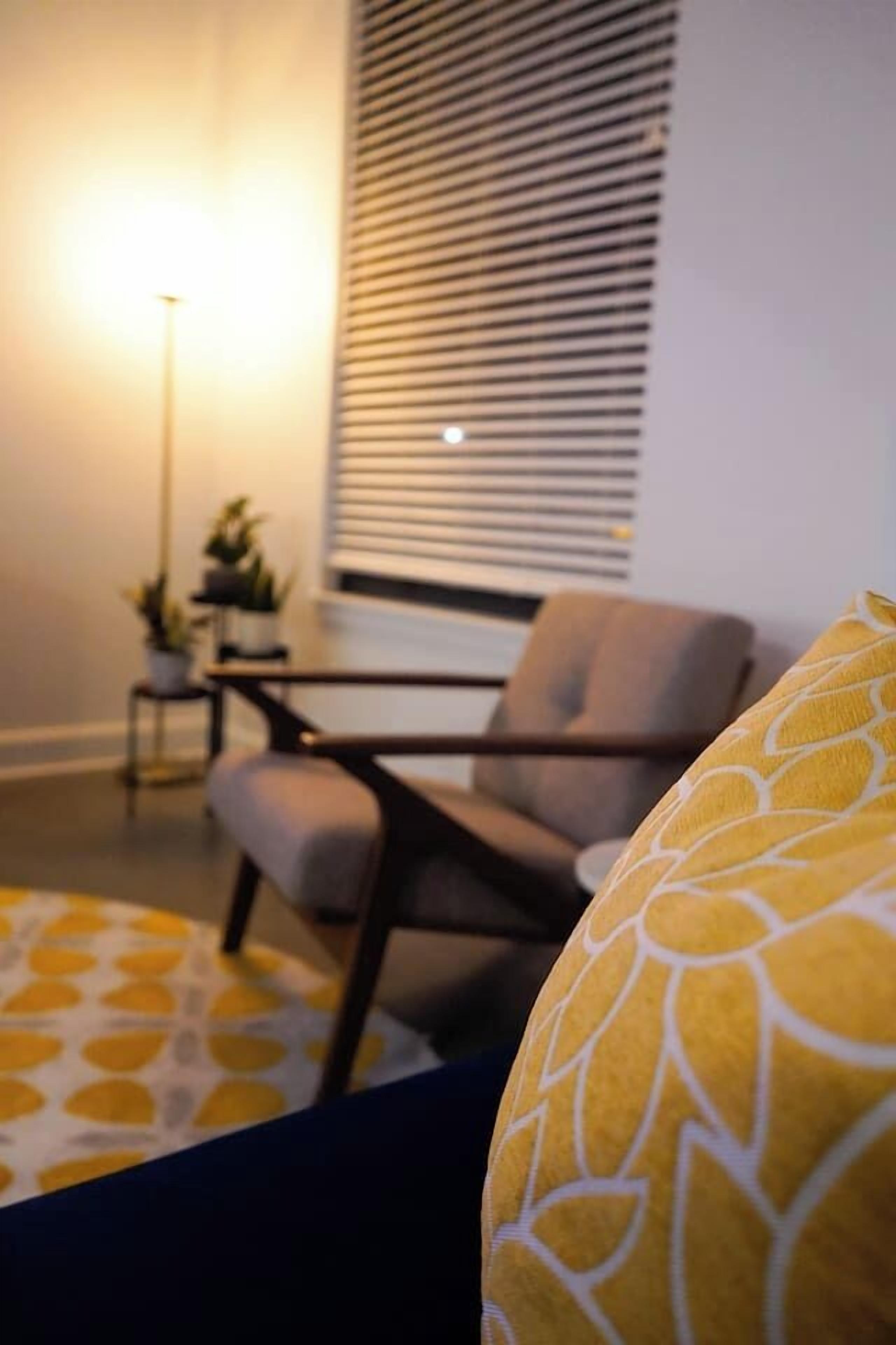Ein Zimmer mit einem modernen Sessel, einem kleinen Tisch mit Pflanze und einem Fenster mit Jalousien.