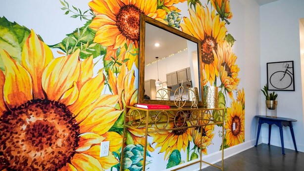 A room with a mural of sunflowers on the wall, a mirror, and a small table with a plant.