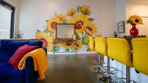 A room with a blue sofa, yellow chairs, a mirror, and a sunflower mural.