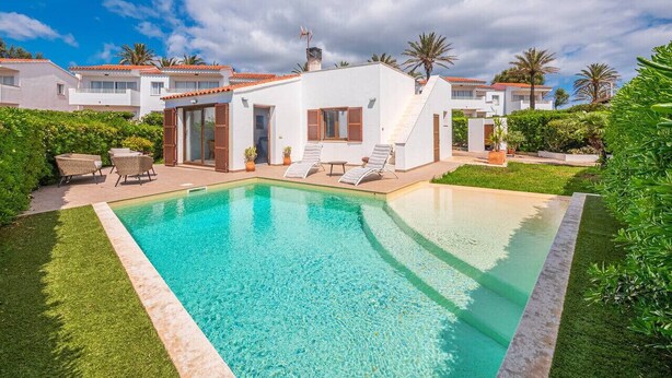Una piscina con una casa blanca al fondo, rodeada de follaje y palmeras.