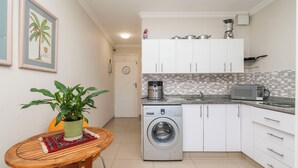 A compact kitchen with white cabinets, a washing machine, a microwave, and a small dining table with a plant. - CapeCity Studio~ Walk 5mins>V&A Waterfront & CTICC (Cape Town)