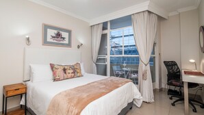 A bedroom with a large bed, a desk, and a chair. There is a window with curtains, a picture on the wall, and a small table beside the bed. - CapeCity Studio~ Walk 5mins>V&A Waterfront & CTICC (Cape Town)