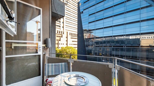 A balcony with a glass table and chair, a white door, and a view of modern buildings. - CapeCity Studio~ Walk 5mins>V&A Waterfront & CTICC (Cape Town)