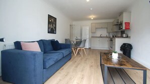 A compact living space with a blue sofa, a dining table, and a kitchenette in the background. - NEW Immaculate Garden Flat (Cliftonville)