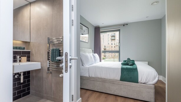 A modern hotel room with a bed, a built-in shower, a window with a view of buildings, and a small shelf with towels.