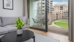 A modern living room with a grey sofa, a round black table, and a potted plant. A sliding glass door leads to an outdoor balcony with a view of a residential building and a green lawn.