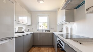 A modern kitchen with grey cabinets, a wooden floor, a window, and a stainless steel range hood. - Kate Kennedy Court (No 2) (St Andrews)