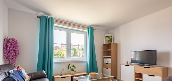 A living room with a sofa, a glass coffee table, a TV on a wooden stand, a bookshelf, and a window with teal curtains. - Kate Kennedy Court (No 2) (St Andrews)